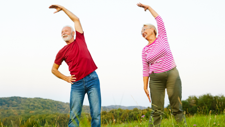 Ein älteres Paar führt Dehnübungen im Freien auf einer Wiese durch, umgeben von Landschaft und Hügeln.