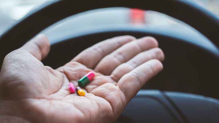 Eine Hand hält bunte Kapseln über dem Lenkrad eines Fahrzeugs.