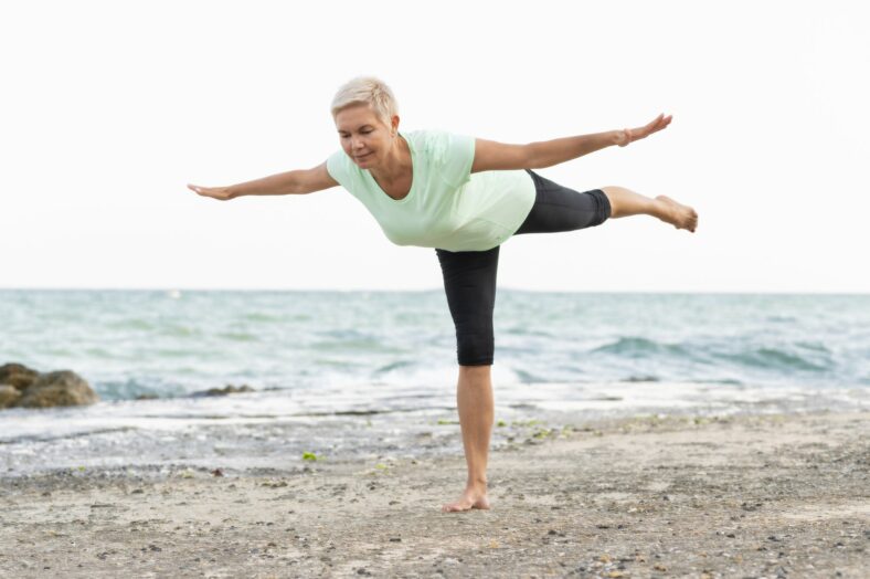 Eine Frau in einem hellgrünen Shirt und schwarzen Leggings balanciert barfuß am Strand, mit Blick auf das Wasser.