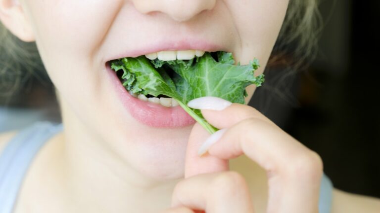 Eine Person mit blonden Haaren hält ein Blatt Grünkohl vor ihren Mund, bereit zum Essen.