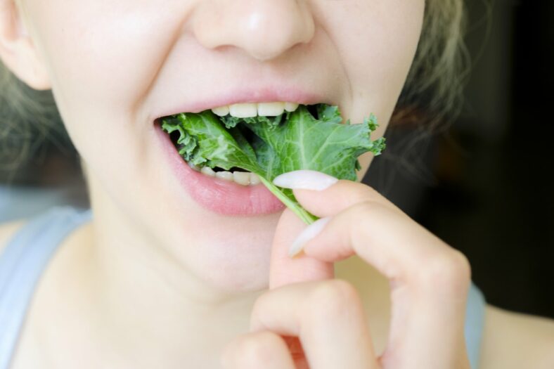 Eine Person mit blonden Haaren hält ein Blatt Grünkohl vor ihren Mund, bereit zum Essen.