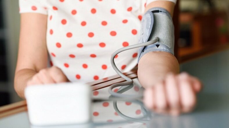 Eine Person mit einem Blutdruckmessgerät am Arm, das auf einem Tisch liegt. Im Hintergrund ein unscharfer Raum.
