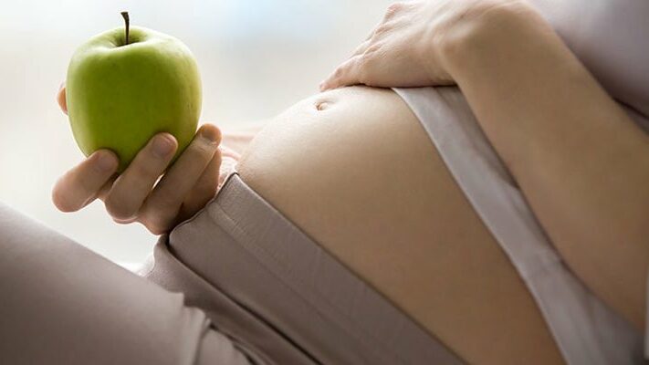 Eine schwangere Person hält einen grünen Apfel und berührt sanft ihren Bauch.