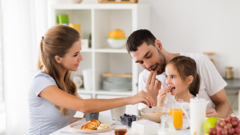 Familie beim gemeinsamen Frühstück an einem Tisch mit Obst, Pfannkuchen und Getränken.