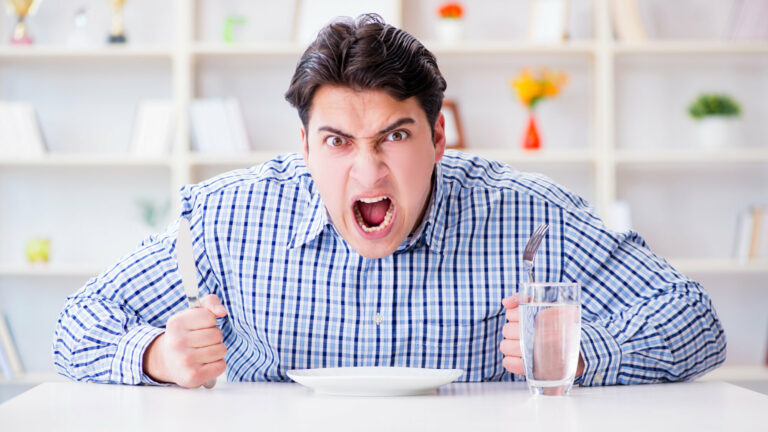 Ein wütender Mann hält ein Messer und eine Gabel über einem leeren Teller, während er an einem Tisch sitzt.