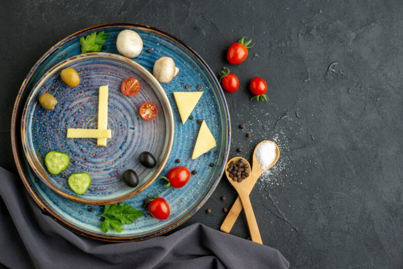 Ein kreativ angerichteter Teller mit Gemüse, Käse und Gewürzen in Form einer Uhr auf blauem Geschirr.