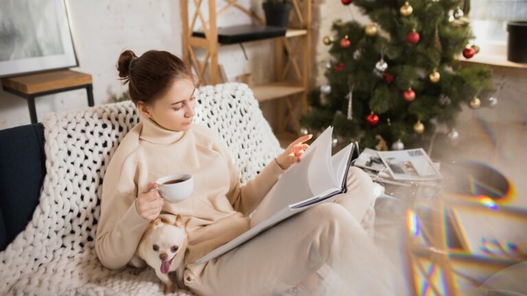 Eine Frau in bequemer Kleidung sitzt auf einem Sofa, liest in einem Buch und hält eine Tasse Kaffee, während ein Hund bei ihr sitzt.