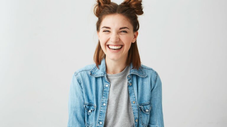 Lachende junge Frau mit Duttfrisur, trägt eine Jeansjacke über einem grauen T-Shirt, vor einem hellen Hintergrund.