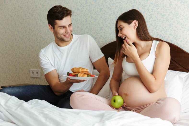 Ein schwangeres Paar sitzt im Bett, der Mann bietet der Frau mit einem Lächeln eine Platte mit Gebäck an, während sie einen Apfel hält.