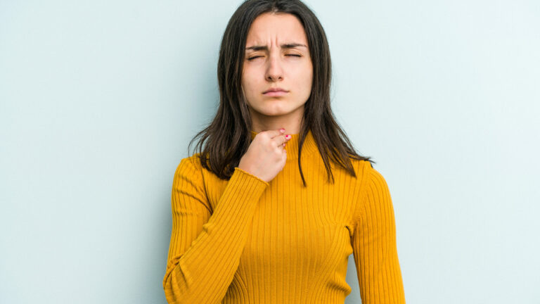 Frau in gelbem Pullover, die nachdenklich auf ihre Kehle zeigt, vor einer hellblauen Wand.
