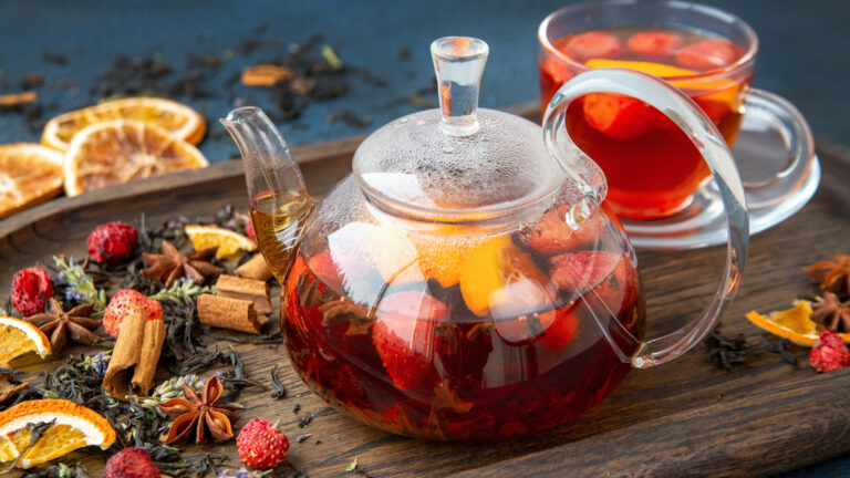 Teekanne aus Glas mit fruchtigem Tee, umgeben von Trockenfrüchten und Gewürzen auf einem Holzbrett.