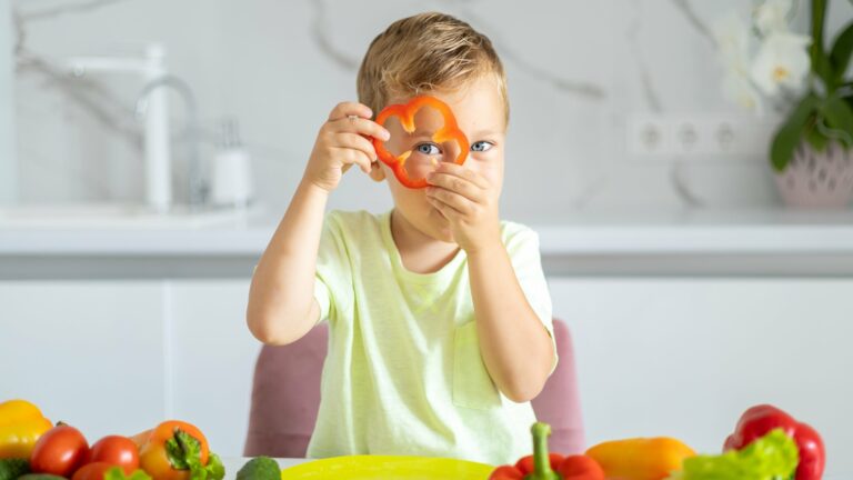 Ein Junge hält eine rote Paprika in Form eines Blumenmotivs und schaut in die Kamera, umgeben von frischem Gemüse.