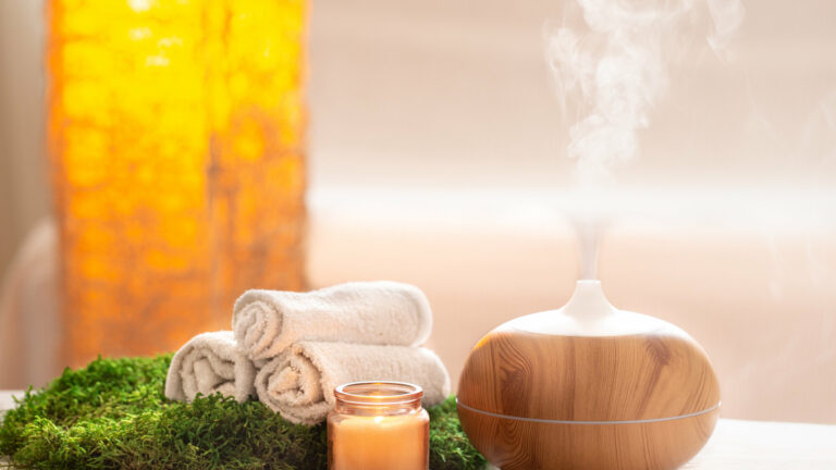 Ein Aromadiffusor aus Holz mit aufsteigendem Dampf, umgeben von Handtüchern, einer Kerze und grünem Moos.