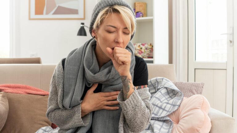 Eine Frau mit grauem Schal und Mütze hustet auf einem Sofa, umgeben von Decken und Kissen.