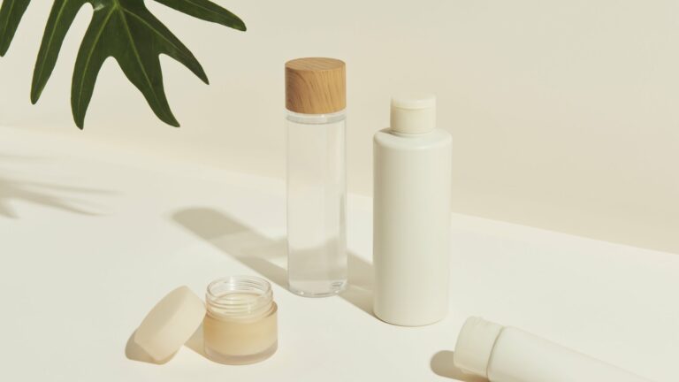 Verschiedene kosmetische Flaschen und Tiegel, darunter ein Glas mit Holzstoß, auf einem hellen Untergrund.