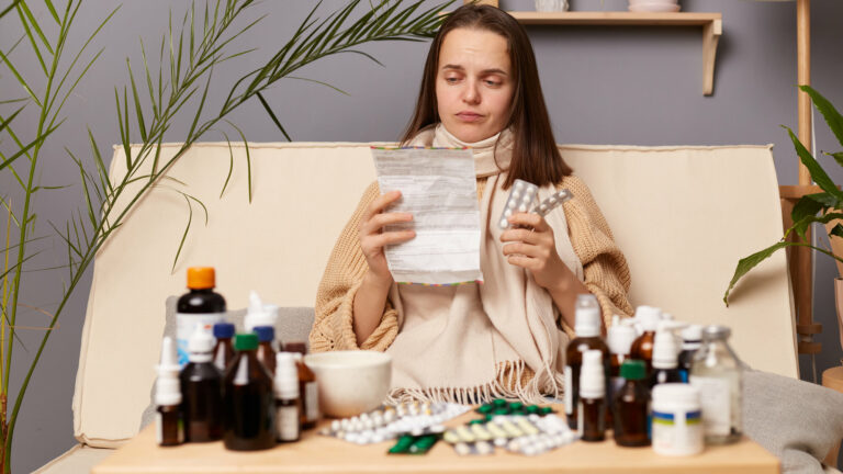 Eine kranke Frau sitzt auf einem Sofa, umgeben von verschiedenen Medikamenten und hält einen Zettel sowie eine Medikamentenpackung in der Hand.