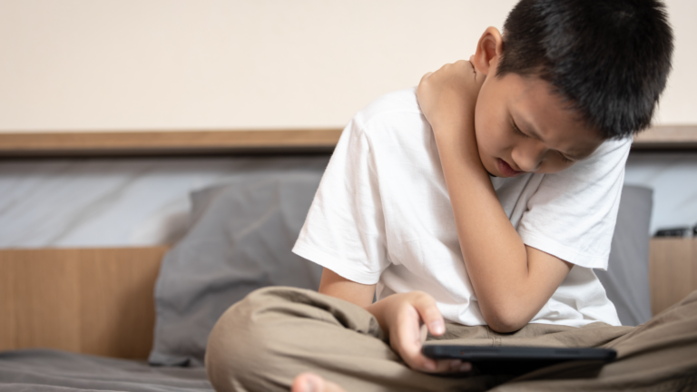 Ein Junge sitzt auf einem Bett, hält ein Tablet und fasst sich schmerzverzerrt an den Hals.